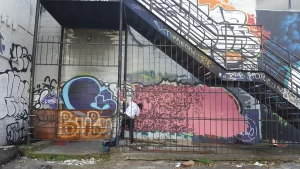 A man painting graffiti on a building under a set of stairs in Houston, Texas on a rainy day.