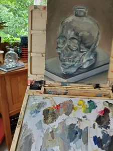 An oil painting of a crystal skull sits on an easel with mixtures of used oil paint on the palette. In the background sitting on a table is that same crystal skull which has been painted.