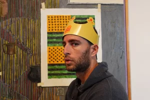 A man wearing a burger king paper crown posing for a photo in front of a jasper johns painting titled ventriloquist at the Houston museum of fine arts