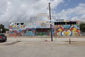 The East facing side of Houston Graffiti Park with clouds and bright blue sky. The walls are brightly painted with different styles and colorful murals decorating the facade.