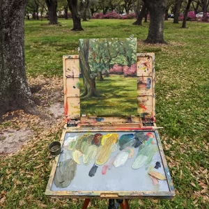 A painting of a landscape with trees and bushes sits on a plein air painting easel. the oil painting appears to have just been finished because many colors such as greens and yellows still remain on the palette.