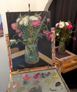 An oil painting of a floral arrangement sits on an easel with paint on the pallet while in the background that same floral arrangement sits on a table.