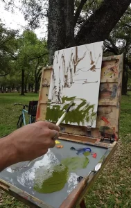 a hand holding a paint brush applies green paint of a white sheet of paper that is attached to a plein air painting easel. The location of the oil painting is in houston texas with trees in the background of a grey day.